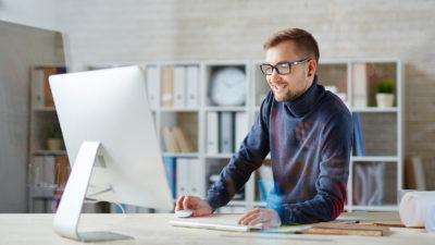 Hyvin pukeutunut mies istuu toimistossa tietokoneen edessä, katsoo näyttöä ja hymyilee